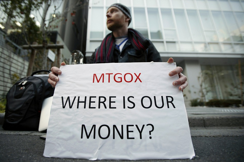 A man sitting on a sidewalk in front of a glass building holding a white sign that reads &ldquo;MTGOX WHERE IS OUR MONEY?&rdquo; in black and red letters.